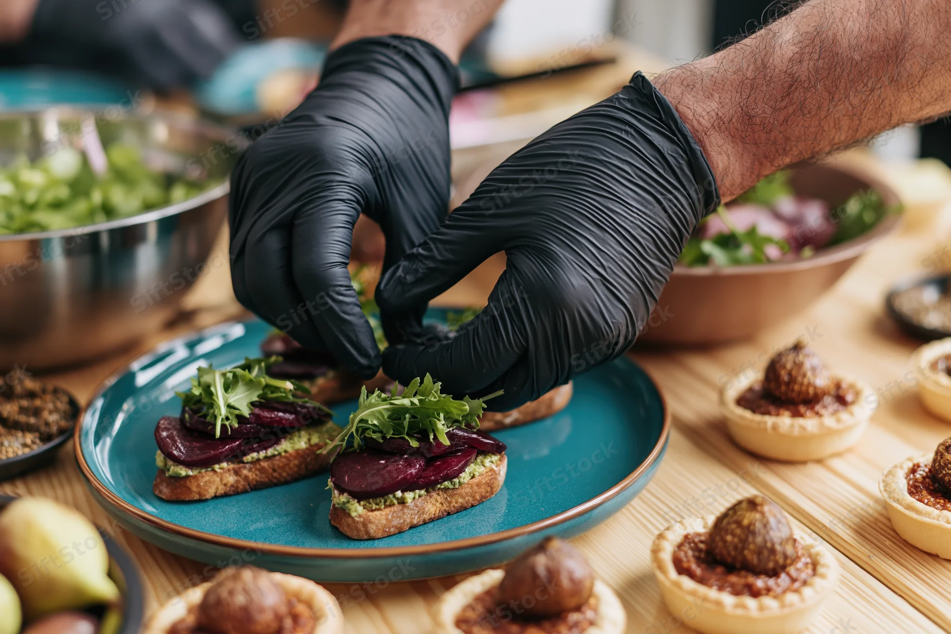 Chef's hands in black nitrile gloves arranging arugula on beet toast with avocado spread on a teal plate surrounded by small tarts and bowls of salad on a wooden table