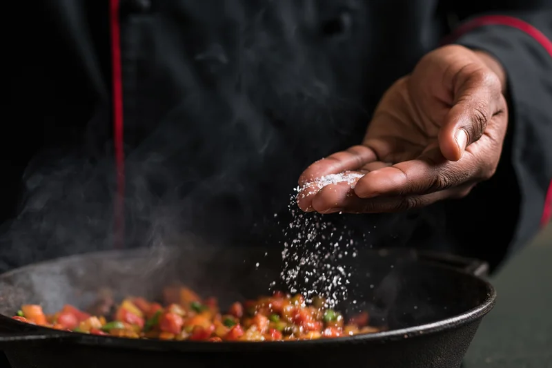 Chef sprinkling salt over steaming sauteed vegetables in a cast iron pan