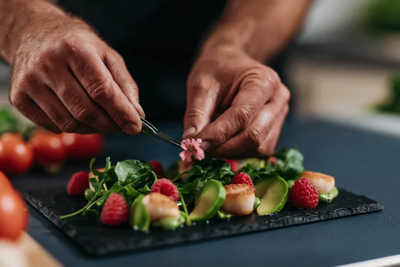 Chef's hands using tweezers to place a pink edible flower on a plated salad with scallops, raspberries, and avocado
