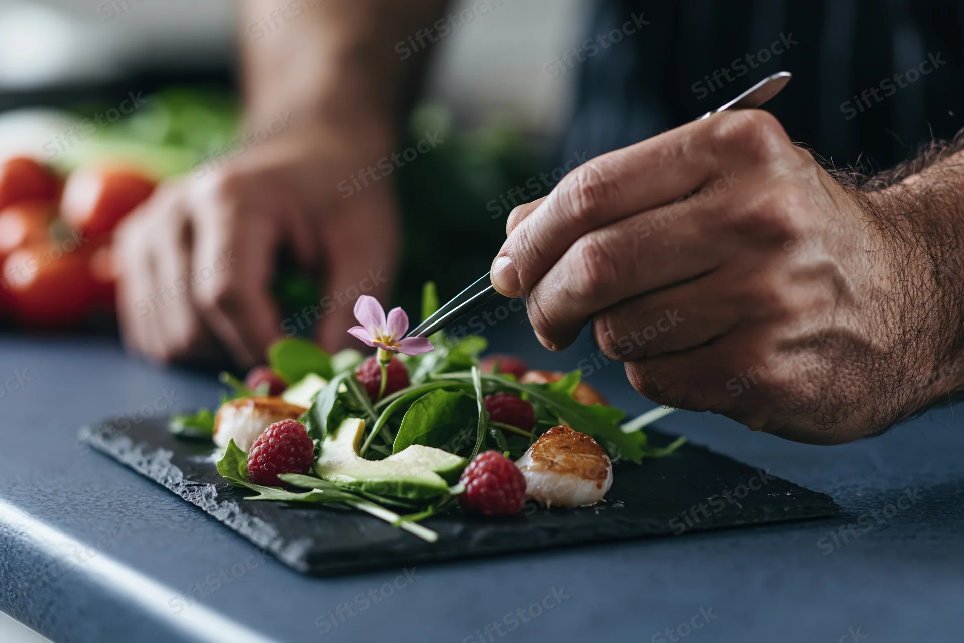 Chef using tweezers to place an edible flower on a salad with scallops and raspberries on a slate plate