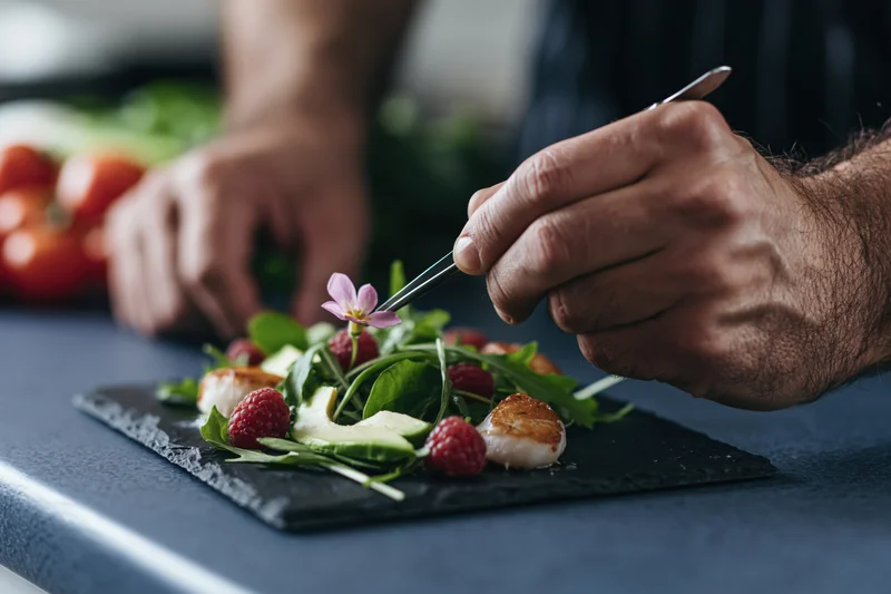 Chef using tweezers to place an edible flower on a salad with scallops and raspberries on a slate plate