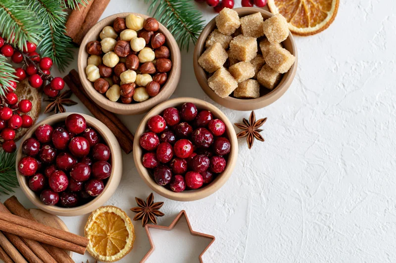 Christmas baking ingredients arranged in wooden bowls with cranberries, hazelnuts, brown sugar, cinnamon, and star anise on a white surface