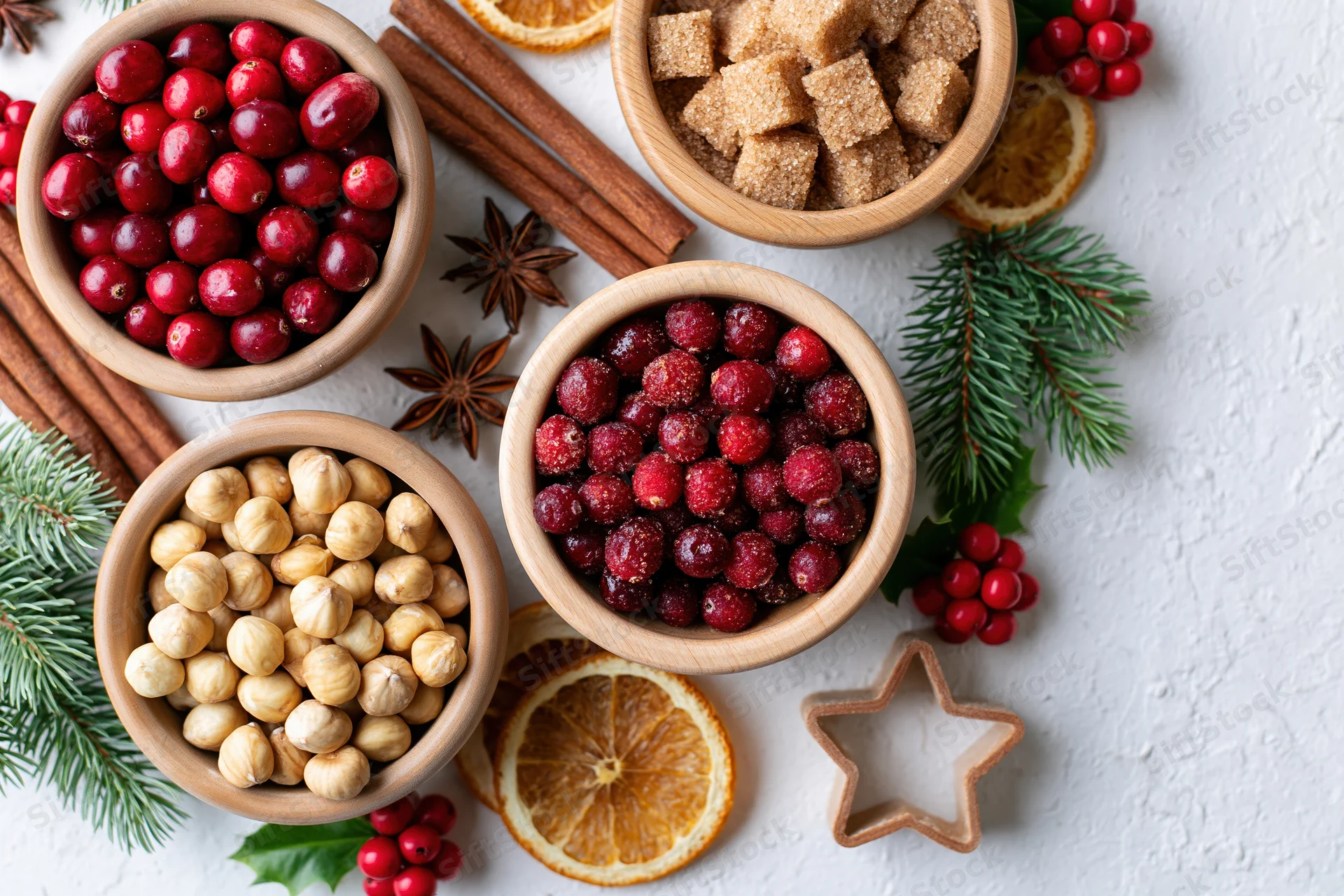 Christmas baking ingredients arranged as a flat lay on a white surface with cranberries, hazelnuts, cinnamon, and evergreen sprigs