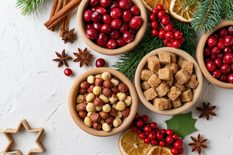 Christmas baking ingredients arranged in wooden bowls with cranberries, hazelnuts, brown sugar, cinnamon, and holly on white surface