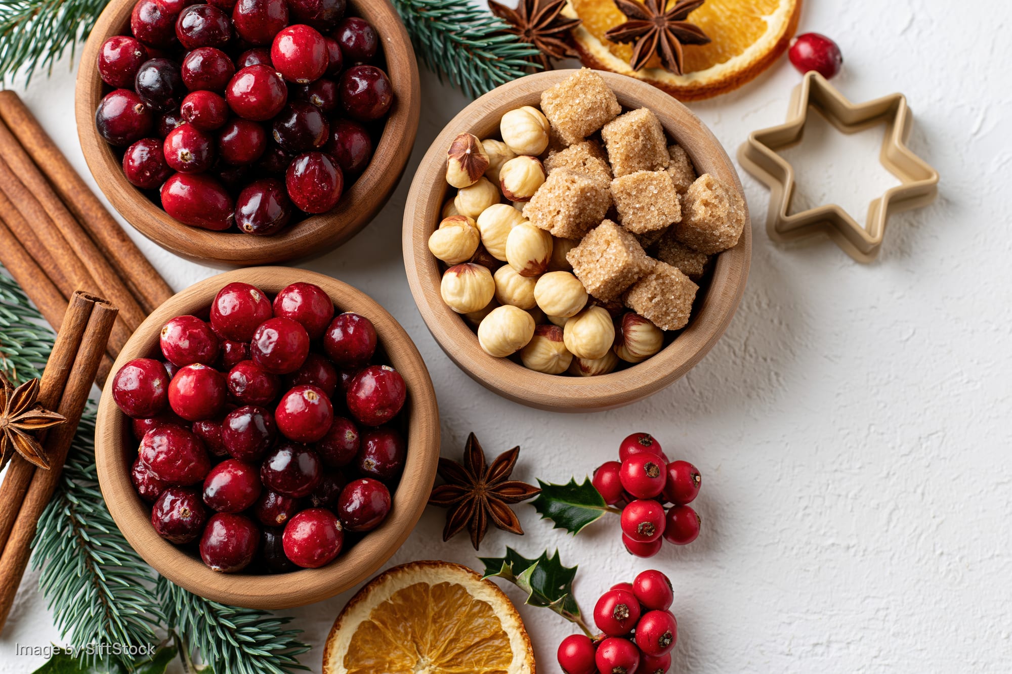 Christmas baking ingredients arranged as a flat lay on a white surface with cranberries, cinnamon, orange, holly, and cookie cutter