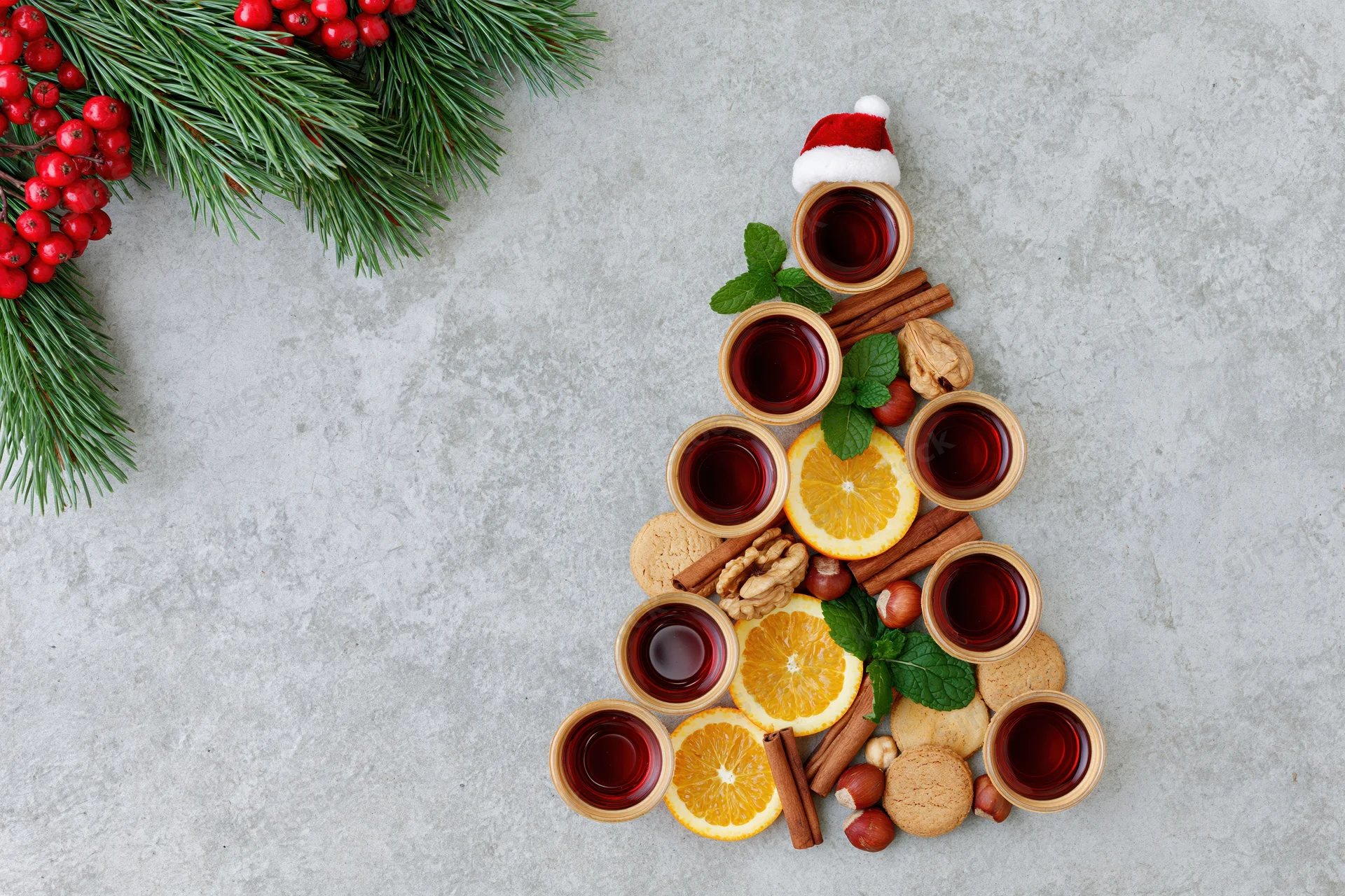 Christmas tree shaped arrangement of mulled wine cups, citrus slices, spices, and cookies on textured grey surface with pine branches and berries
