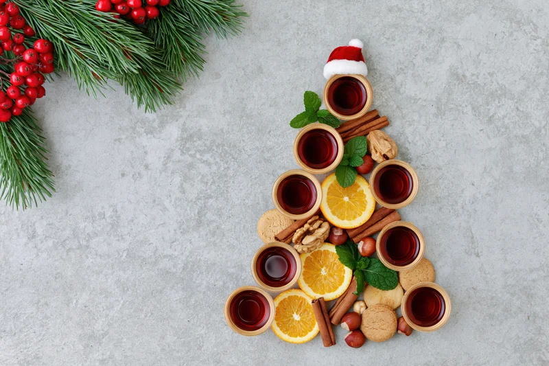 Christmas tree shaped arrangement of mulled wine cups, citrus slices, spices, and cookies on textured grey surface with pine branches and berries