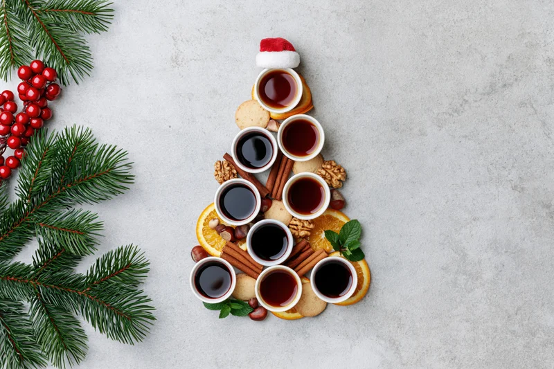 Christmas tree shaped arrangement of tea cups, oranges, cinnamon, nuts and cookies on textured grey surface with pine branches and berries