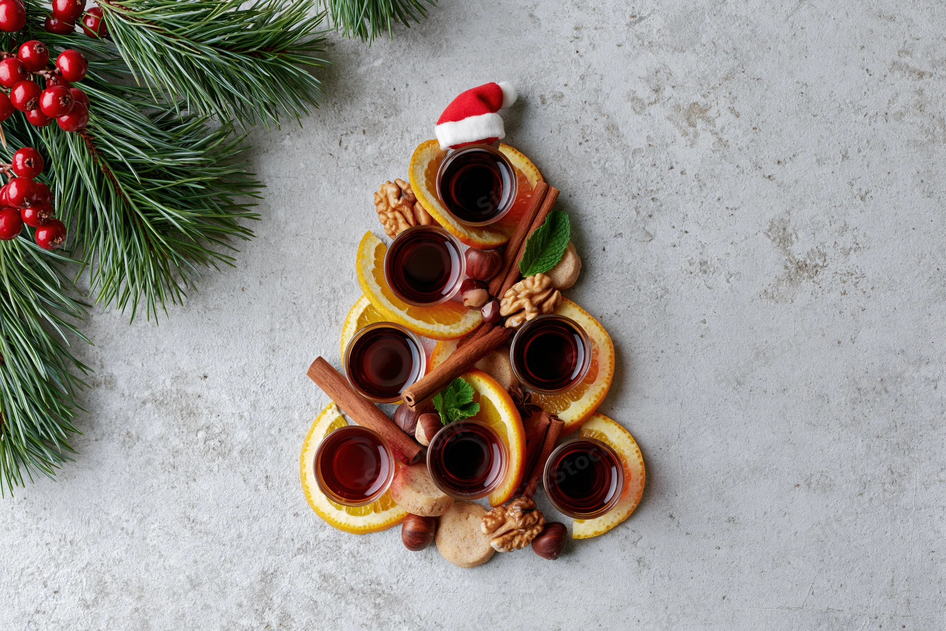 Christmas tree shaped arrangement of orange slices, cinnamon sticks, walnuts, cookies, and mulled wine glasses on textured grey surface with pine branches and berries nearby
