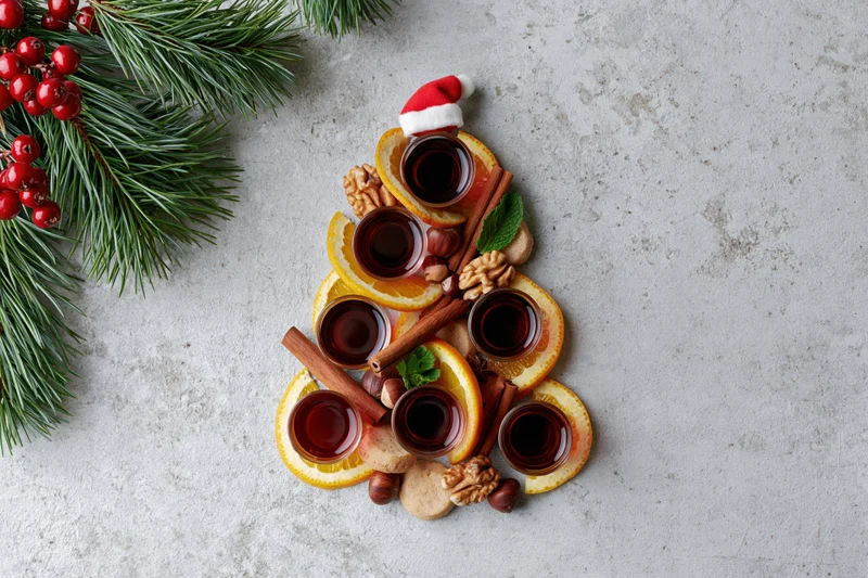 Christmas tree shaped arrangement of orange slices, cinnamon sticks, walnuts, cookies, and mulled wine glasses on textured grey surface with pine branches and berries nearby