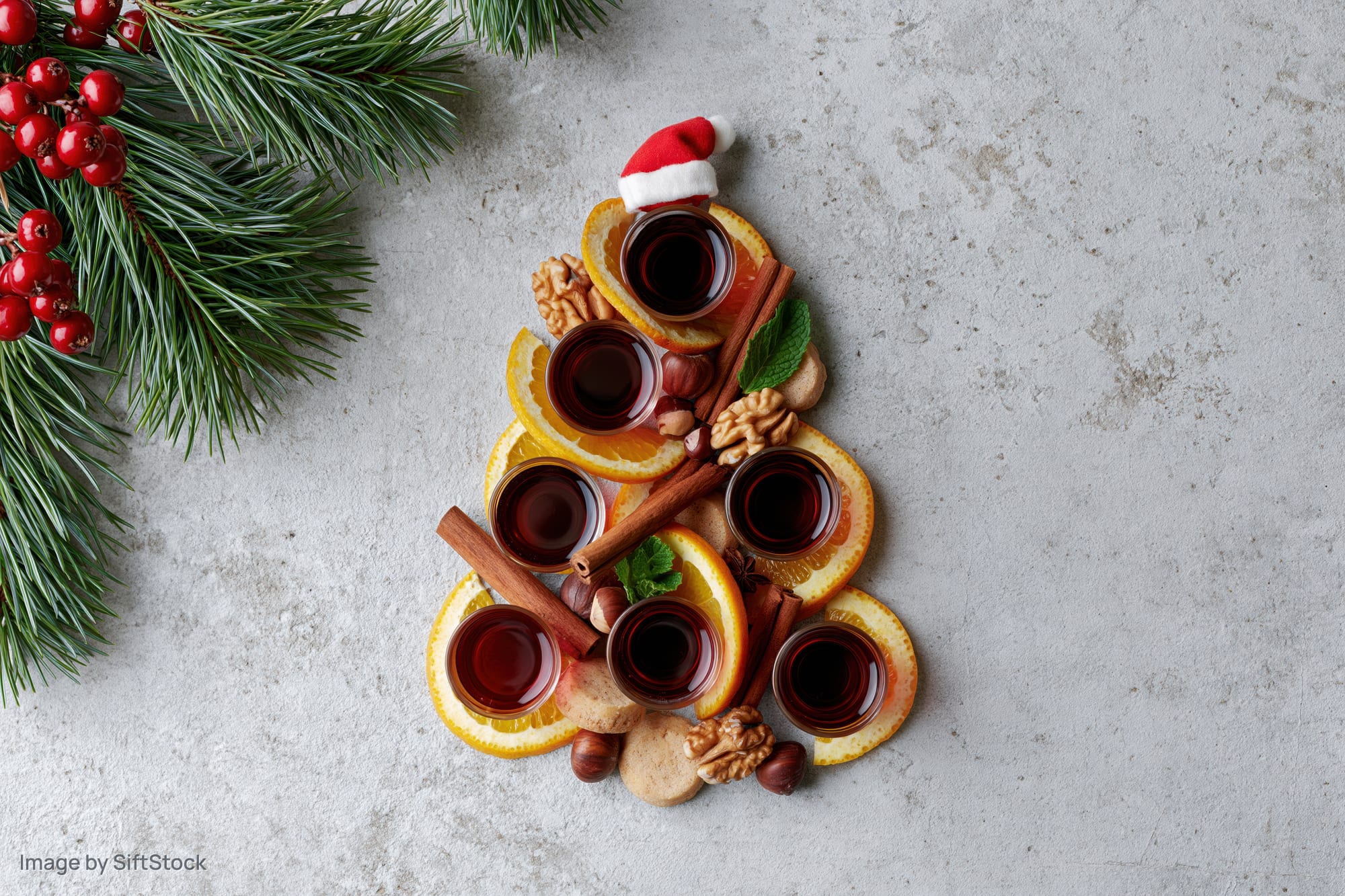 Christmas tree shaped arrangement of orange slices, cinnamon sticks, walnuts, cookies, and mulled wine glasses on textured grey surface with pine branches and berries nearby