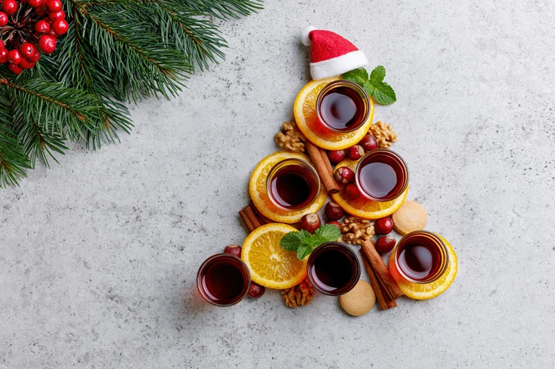 Christmas tree shaped arrangement of oranges, cinnamon, walnuts, mint, berries and mulled wine glasses on textured concrete surface with santa hat and evergreen branch