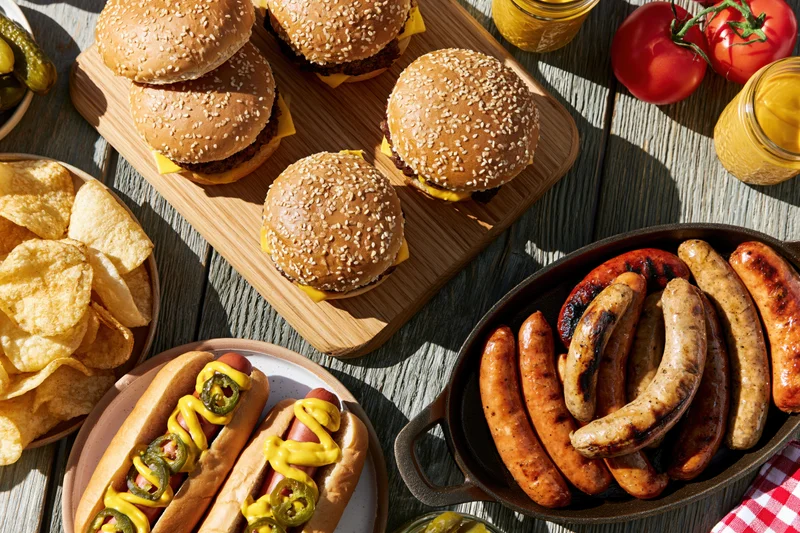 Grilled sausages, burgers, and hotdogs with chips and condiments arranged on a weathered wooden table for a summer barbecue
