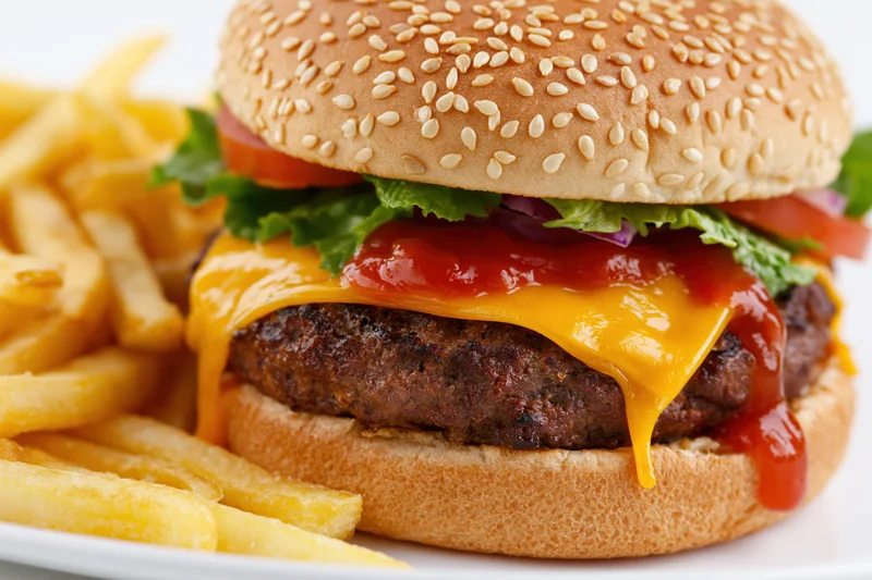 Cheeseburger with sesame seed bun, thick beef patty, melted cheese, ketchup, lettuce, tomato, and onion, served with french fries on a white plate
