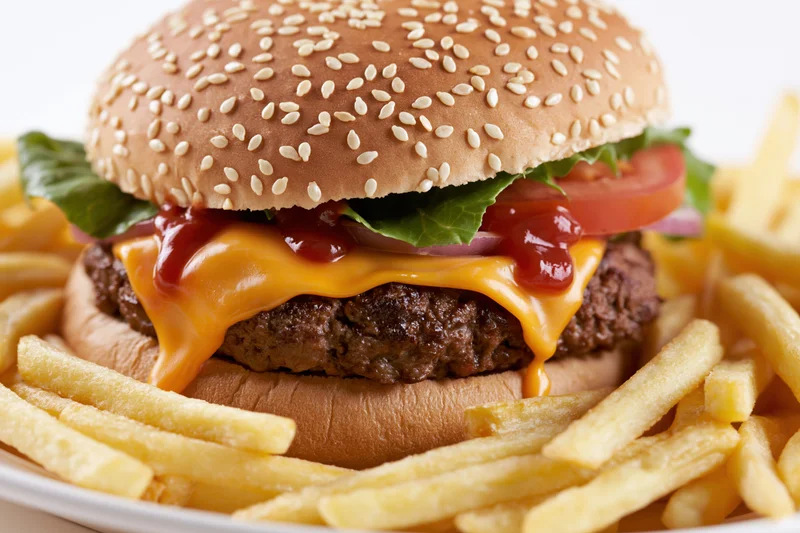 Cheeseburger with sesame seed bun, thick beef patty, melted cheese, ketchup, lettuce, tomato, onion, served with french fries on a white plate
