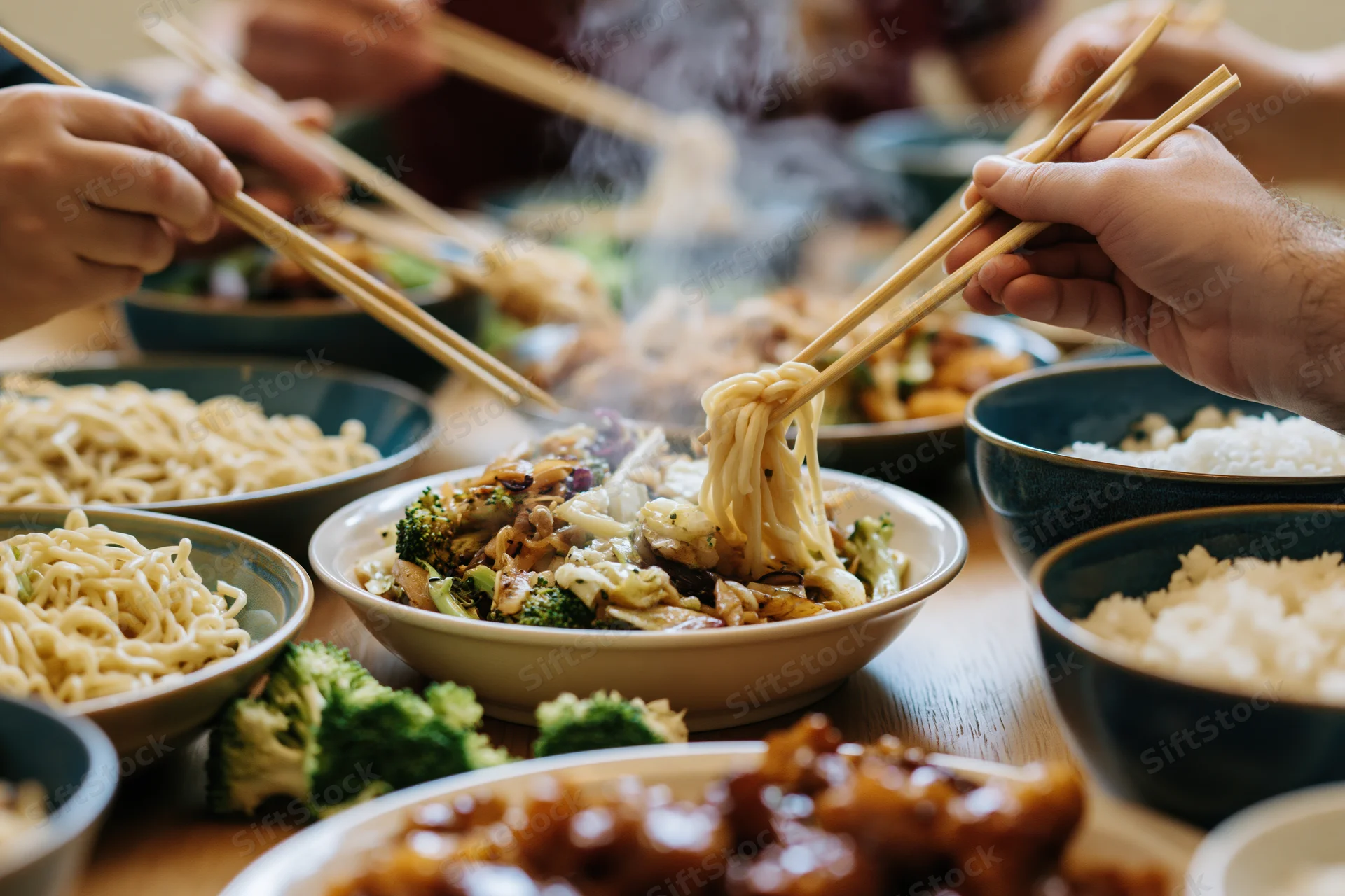 Multiple hands using chopsticks to pick noodles from bowls at a shared dining table