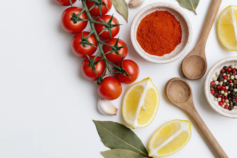 Tomatoes on vine, lemon slices, spices, garlic, bay leaves, wooden spoons, and bowls arranged as a border frame on white surface
