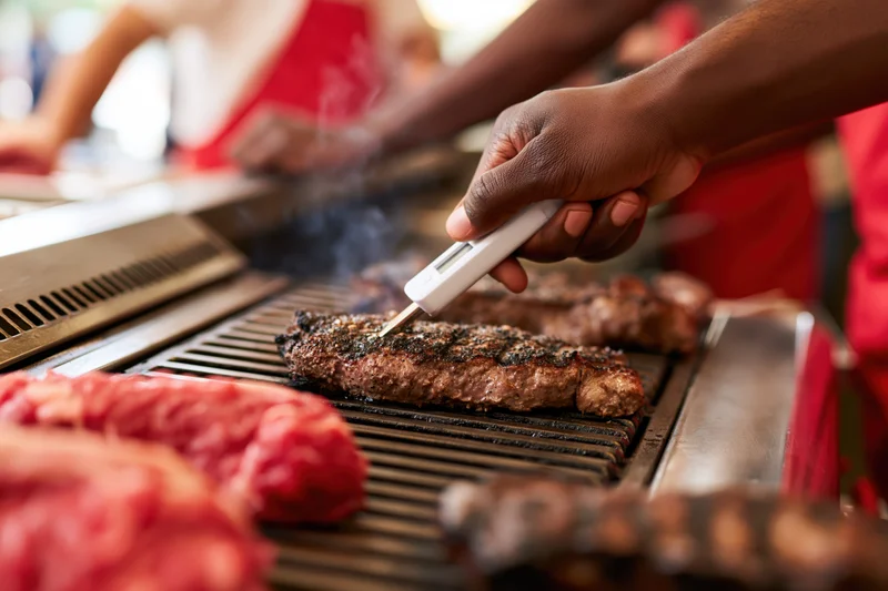 Hand inserting digital meat thermometer probe into grilled steak on barbecue grill