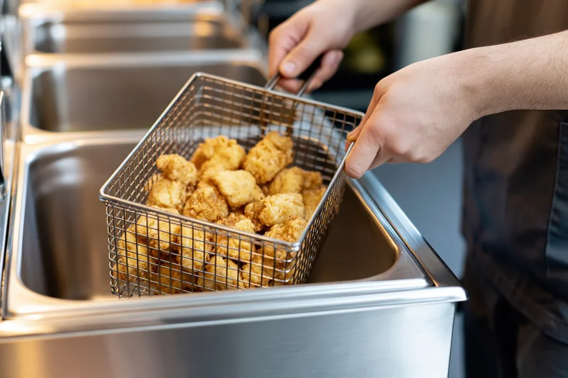 Cook lifting a wire fry basket filled with golden fried chicken in a commercial kitchen