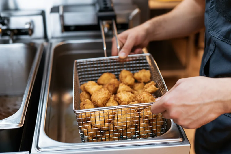 Hands lifting a wire fry basket filled with golden fried chicken from a commercial deep fryer in a restaurant kitchen