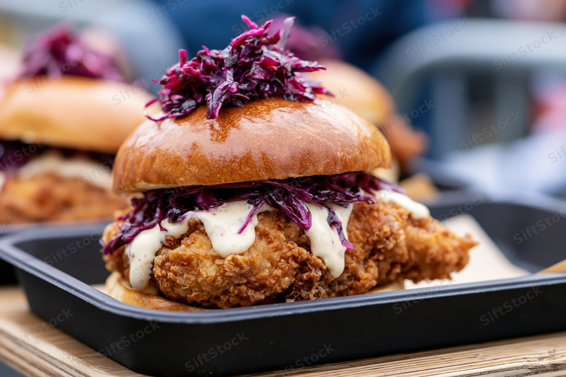 Crispy fried chicken sandwich in a glossy brioche bun with purple cabbage and white sauce on a black tray