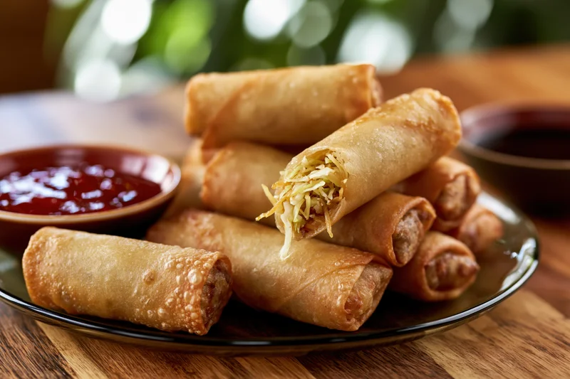 Crispy fried spring rolls piled on a dark glass plate with dipping sauces on a wooden table