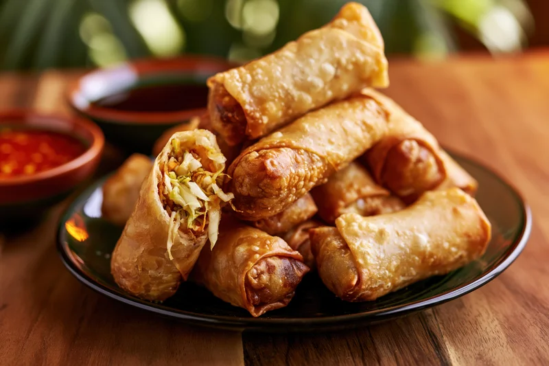 Crispy fried spring rolls piled on a dark glass plate with dipping sauces on a wooden table