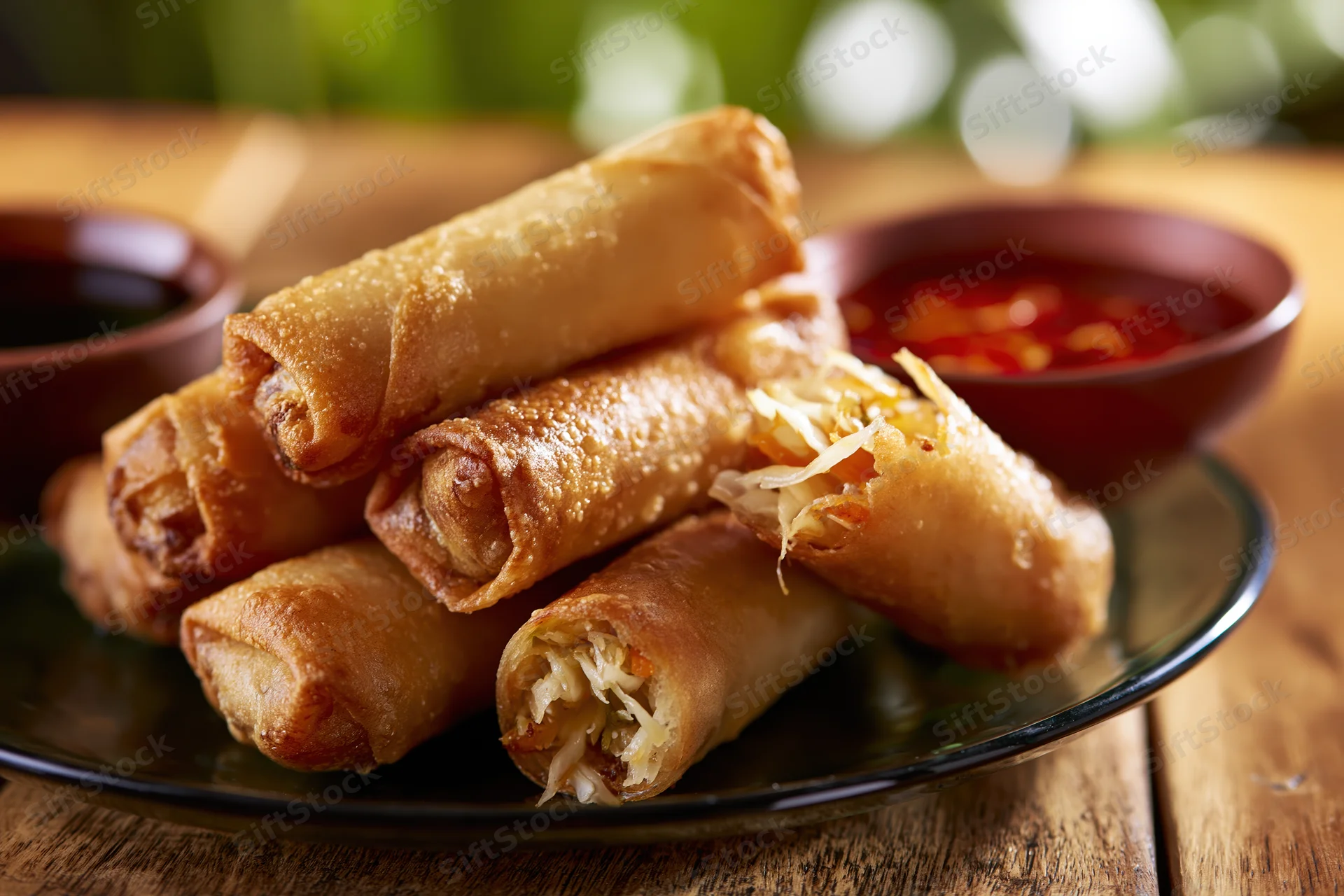 Crispy fried spring rolls piled on a dark glass plate with dipping sauces on a wooden table