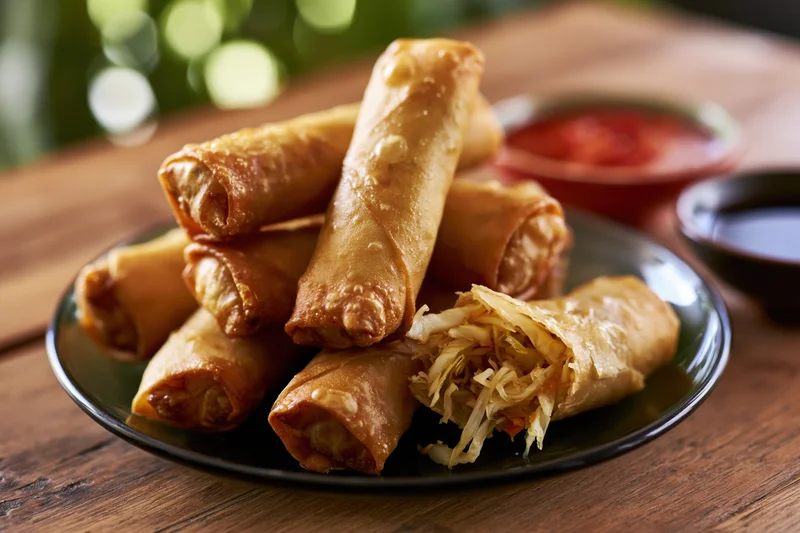 Crispy fried spring rolls piled on a dark glass plate with dipping sauces on a wooden table