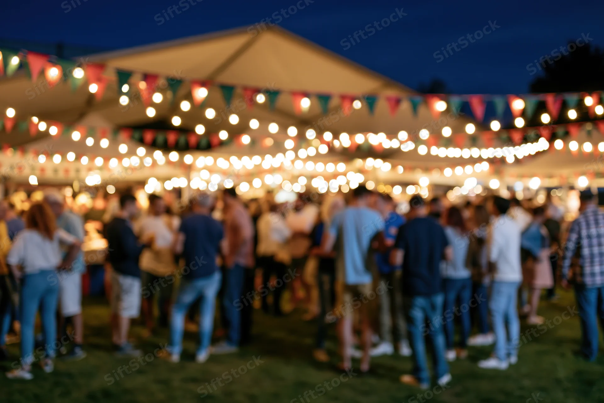 Crowd of people mingling at outdoor night market under string lights and bunting on grassy lawn