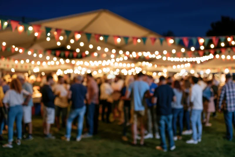 Crowd of people mingling at outdoor night market under string lights and bunting on grassy lawn