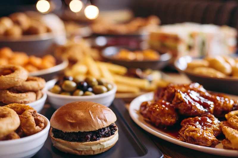 Burger on a table surrounded by chicken wings, onion rings, fries, and olives at a restaurant dinner spread