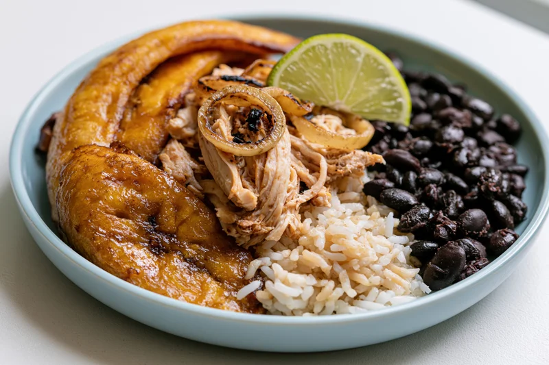 Cuban plate with fried sweet plantains, white rice, black beans, and lime garnish on a light surface