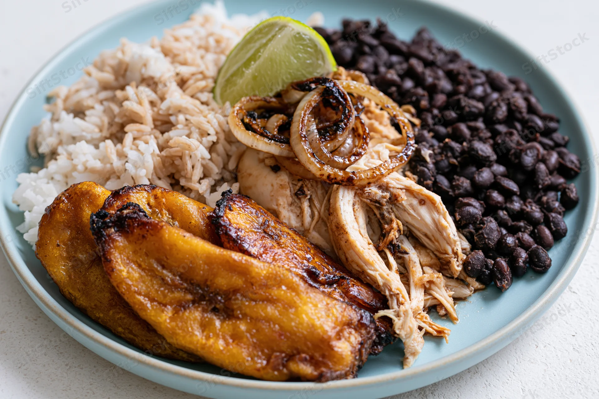 Cuban plate with fried sweet plantains, white rice, black beans, shredded chicken, caramelized onions, and lime garnish on a blue ceramic plate