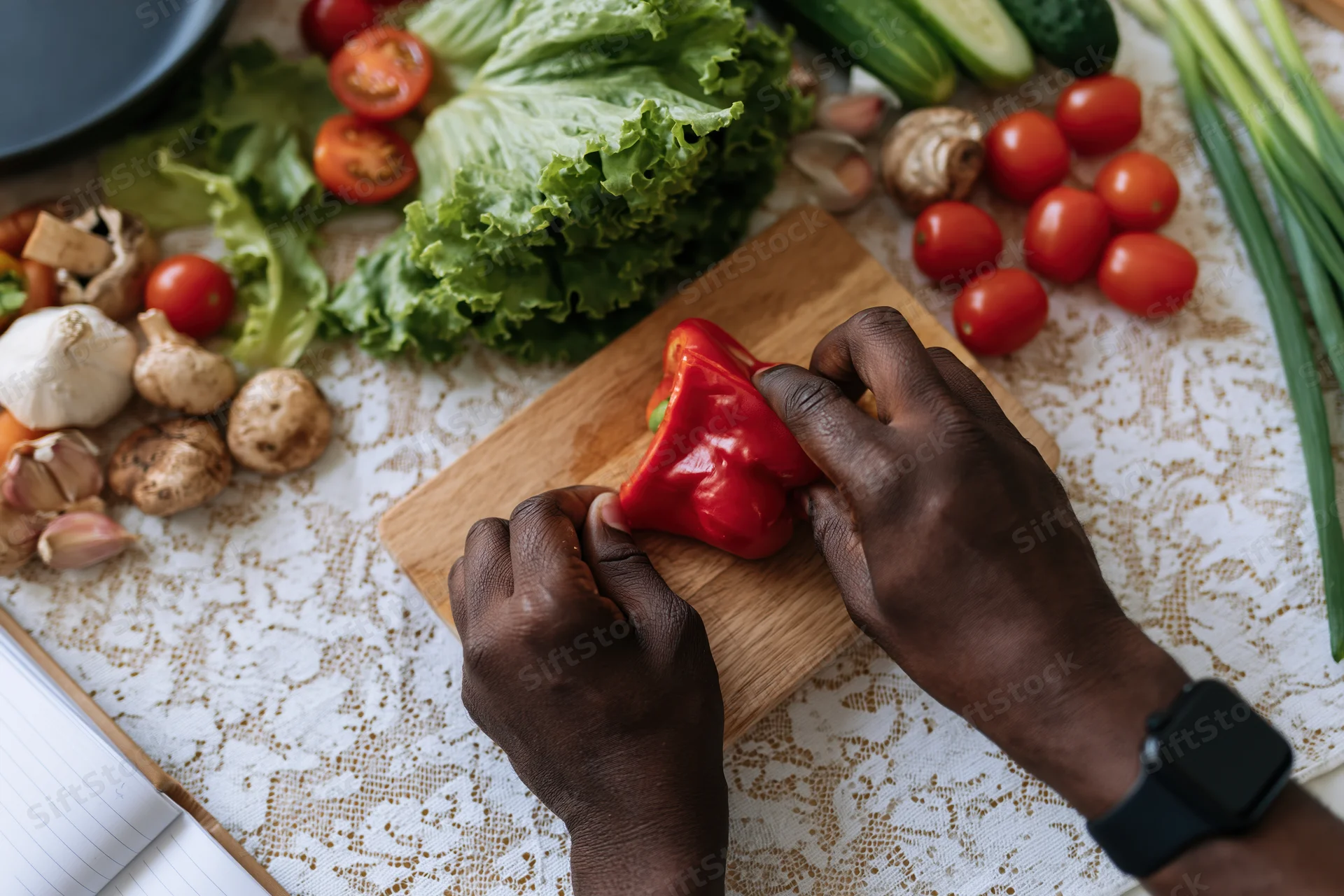 Dark-skinned hands slicing a red bell pepper on a wooden cutting board surrounded by fresh vegetables on a patterned tablecloth