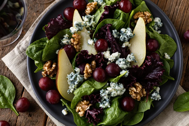 Mixed green salad with pear slices, grapes, walnuts and blue cheese on a dark matte plate on a wooden table