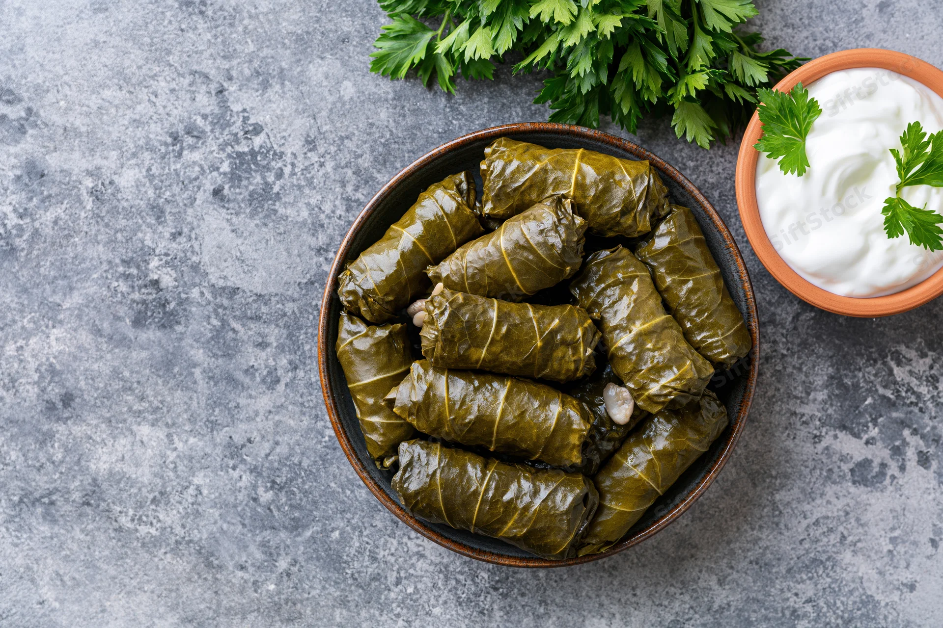 Dolma stuffed grape leaves in a dark ceramic bowl with parsley and yogurt on a gray stone surface