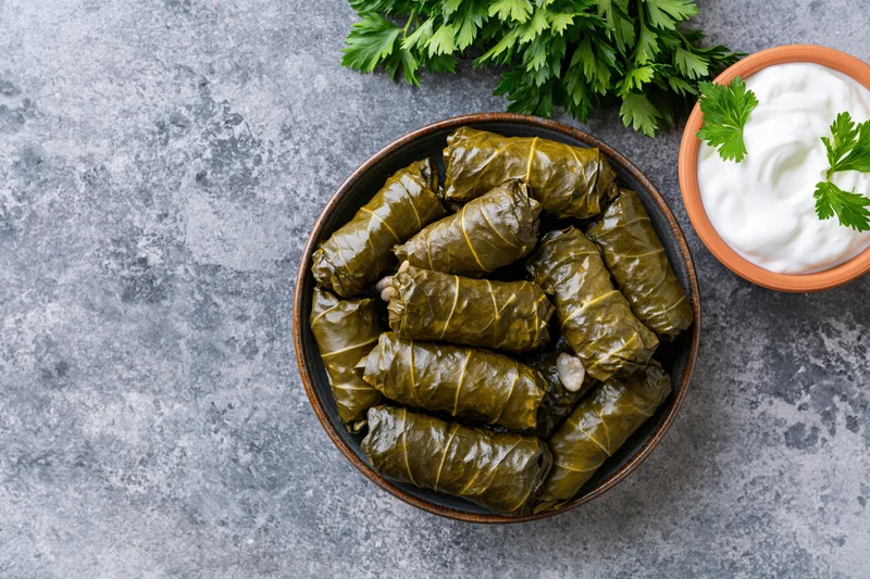 Dolma stuffed grape leaves in a dark ceramic bowl with parsley and yogurt on a gray stone surface