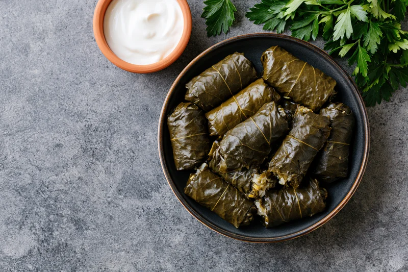 Dolma stuffed grape leaves in a dark ceramic bowl with yogurt dip and parsley on a gray surface