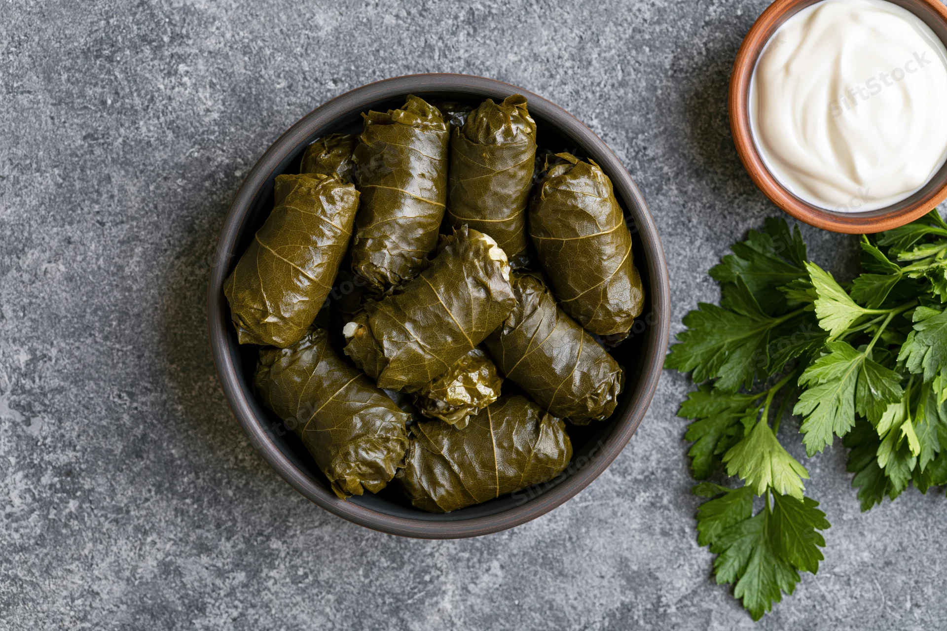 Dolma stuffed grape leaves in a dark ceramic bowl with yogurt and parsley on a gray surface