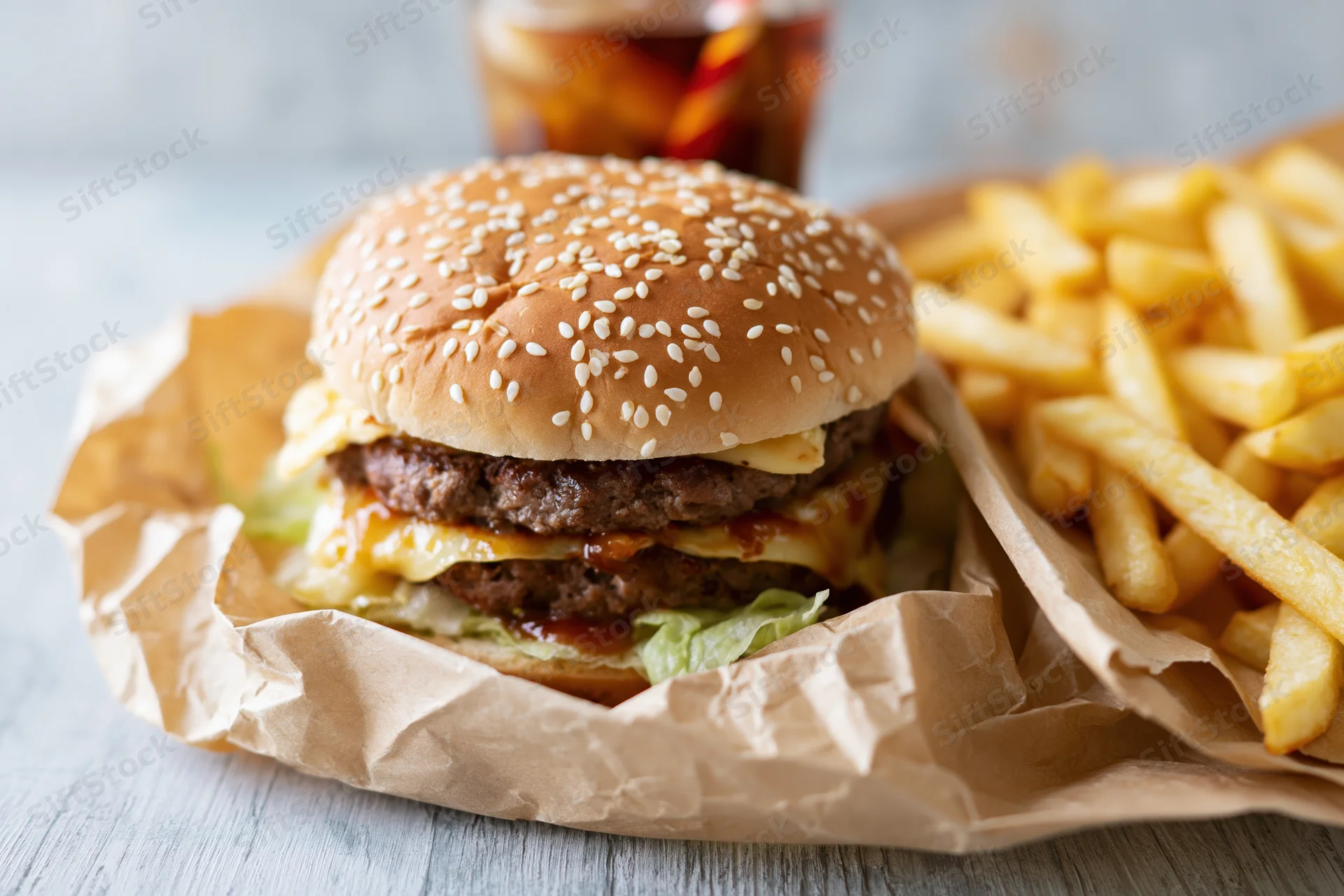 Double-stacked beef burger with cheese and sesame bun served with fries and soda on a wooden table