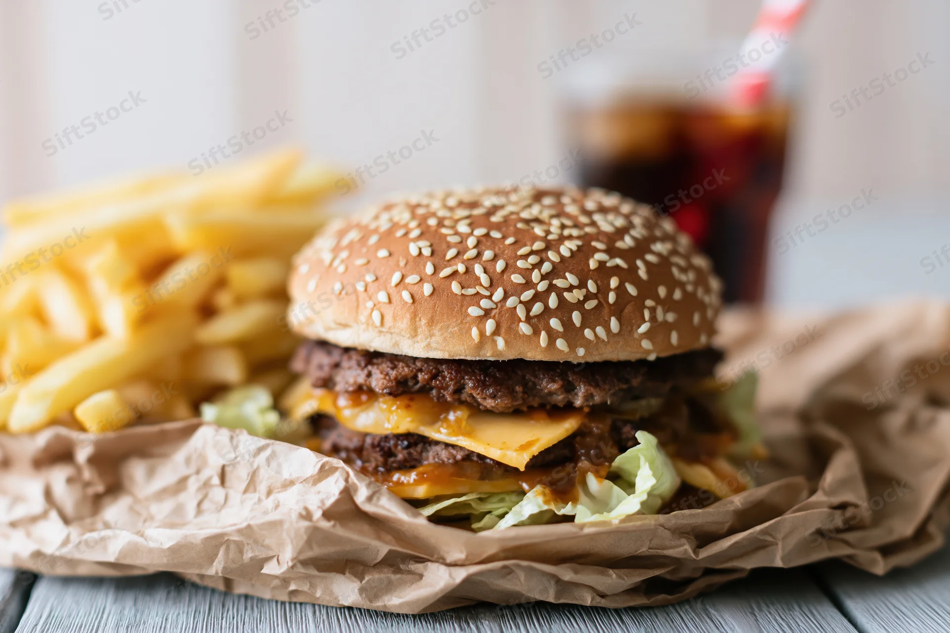 Double-stacked cheeseburger with sesame seed bun, french fries and soda on crumpled paper wrapper on wooden table