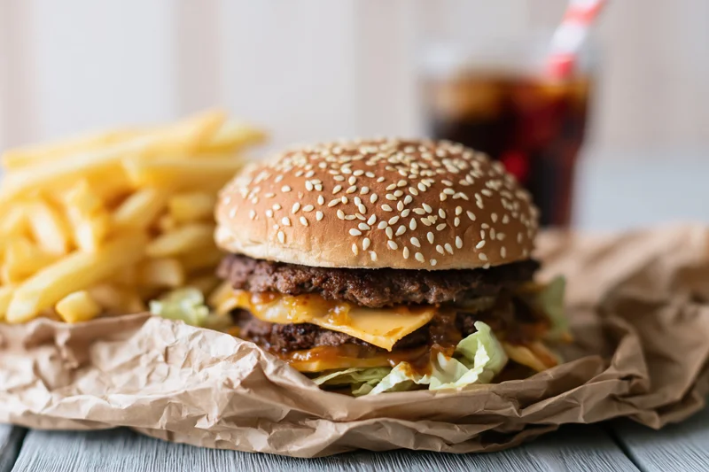 Double-stacked cheeseburger with sesame seed bun, french fries and soda on crumpled paper wrapper on wooden table