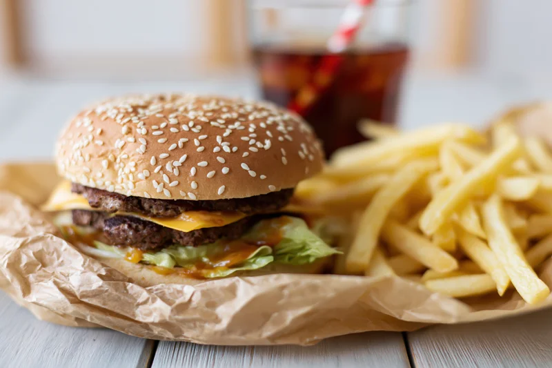 Double-stacked cheeseburger with sesame seed bun, french fries, and soda on a wooden table, served on brown paper wrapper