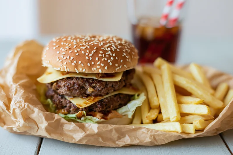 Double-stacked beef burger with cheese and sesame bun served with fries and soda on a paper-lined tray