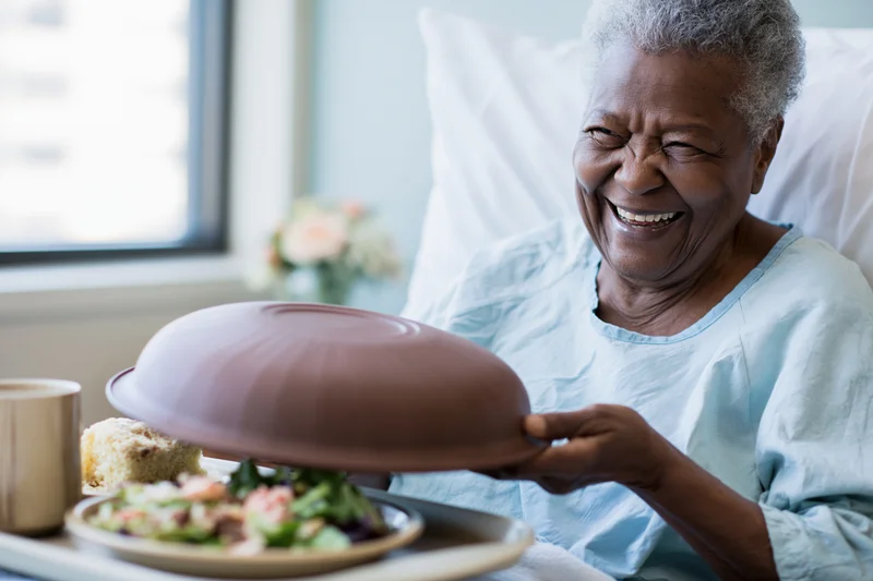 Elderly black woman laughing while lifting lid from meal tray in hospital bed