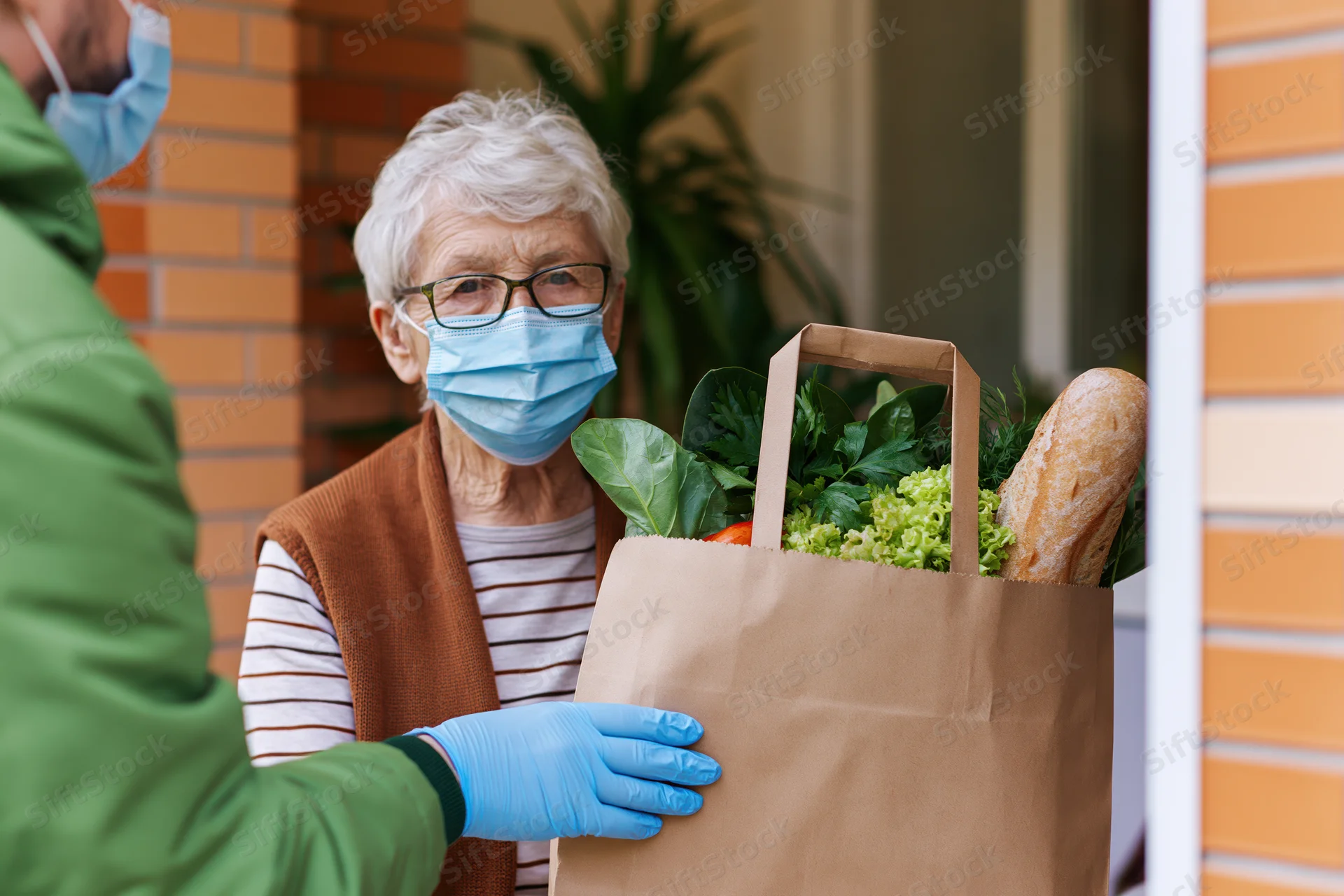 Elderly woman receiving groceries delivery at home porch with protective mask and gloves