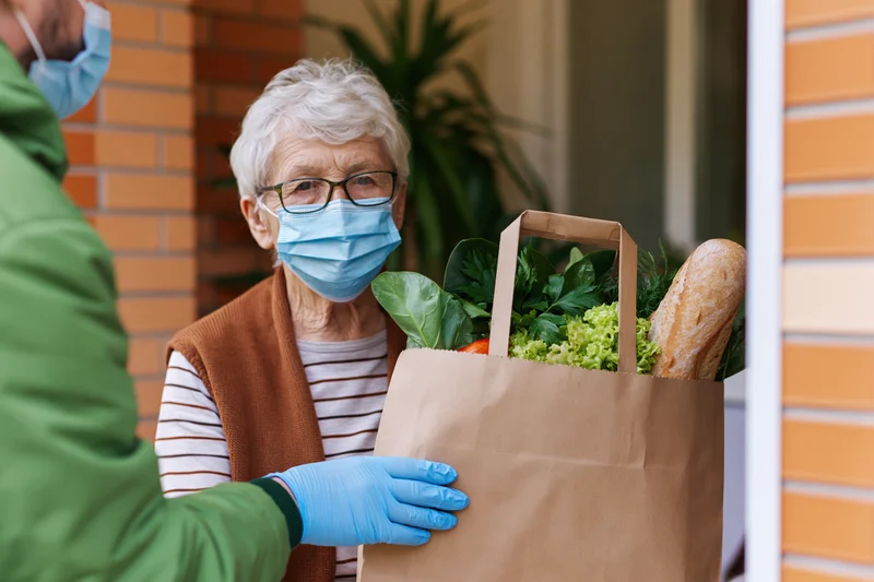 Elderly woman receiving groceries delivery at home porch with protective mask and gloves