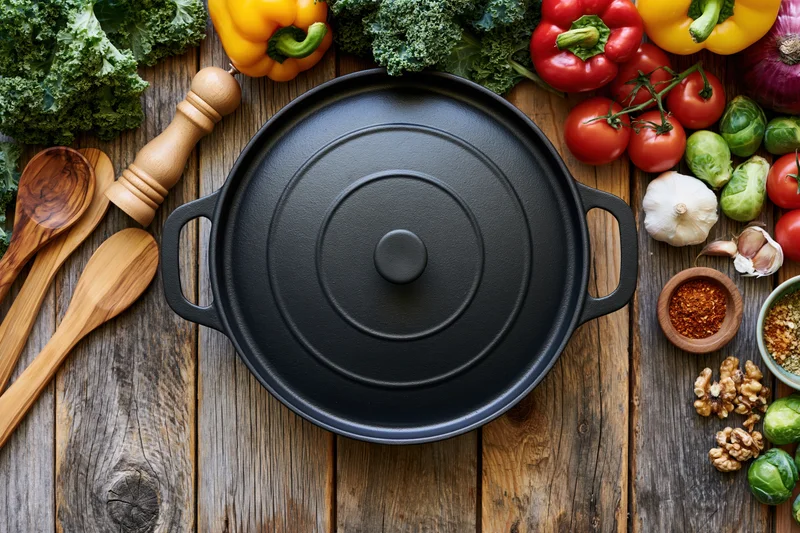 Black cast iron dutch oven centered on weathered wooden table surrounded by fresh vegetables and cooking tools