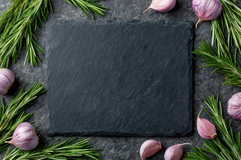 Black slate cutting board centered on dark stone countertop surrounded by rosemary and garlic cloves in flat lay arrangement